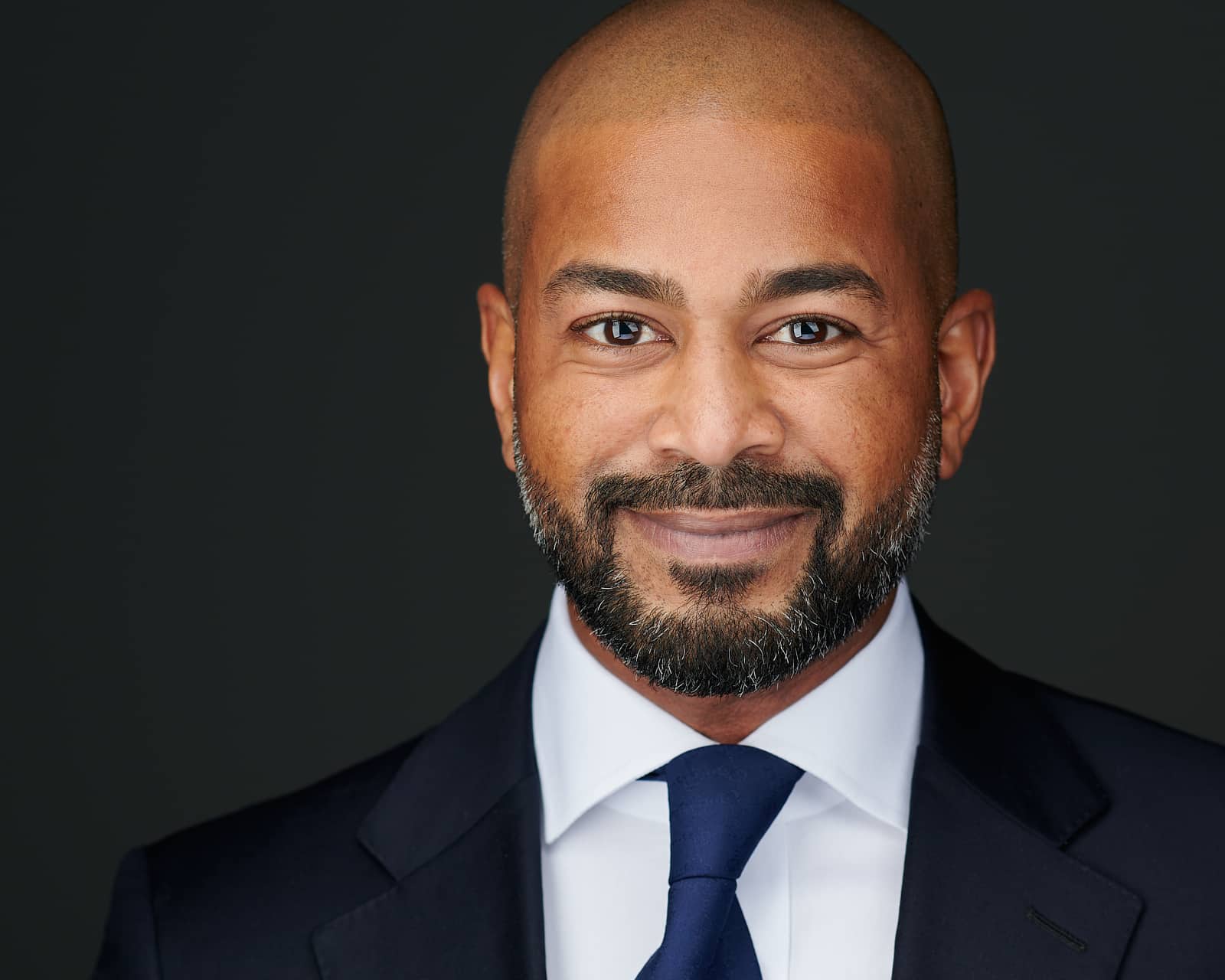 Professional headshot of a successful sales executive, wearing a navy blue suit, a blue tie, and white dress shirt, subtly smiling, against a clean, dark grey background, taken by headshot photographer Heidi Marie Bell in Baltimore Maryland.